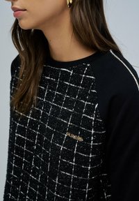 Un pull à carreaux noir et blanc présente un avant texturé et des manches noires unies, rehaussé d'un logo doré sur la poitrine. Encolure arrondie.