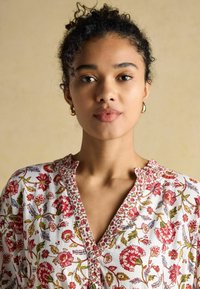 Weiße Bluse mit buntem Blumenmuster in Rot und Grün, mit V-Ausschnitt, kurzen Ärmeln und zarter Borte am Hals und den Knöpfen.