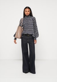 Tengerészes blúz magas nyakkal és puffos ujjakkal, sötét, széles lábú farmerrel párosítva. Egy sima, világosbarna válltáskát visel.