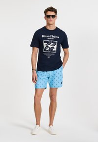 Navy t-shirt with "BlueTides Resort" graphic, paired with light blue swim shorts featuring a cactus pattern. White sneakers and sunglasses.