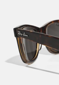 Lunettes de soleil écaille avec une monture épaisse, arborant un logo noir et des verres foncés. Fini lisse et design angulaire.