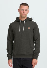 Young man with short hair wearing dark gray hoodie with white drawstrings and front pocket, standing against a plain light background.