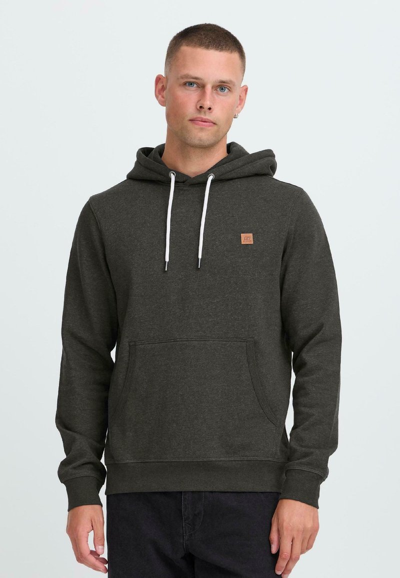 Young man with short hair wearing dark gray hoodie with white drawstrings and front pocket, standing against a plain light background.