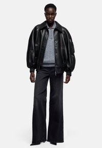 Zwarte leren bomberjack met oversized silhouet, gecombineerd met een grijze trui en wijde zwarte spijkerbroek. Voorzien van ritsen en elastische manchetten.