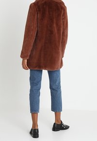 Abrigo de pelaje sintético marrón con cuello, con un corte suelto. El modelo lo combina con unos jeans rectos de color azul claro y zapatos negros de slip-on.