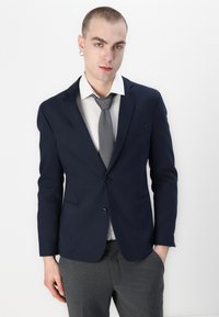 Navy blazer with a tailored fit, single button, and two pockets. Paired with a light shirt and a textured gray tie. Gray tailored trousers.