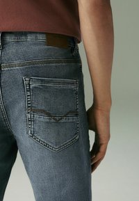 Tmavomodré denimové nohavice so slim fit strihom, ktoré majú výrazný dizajn zadnej vrecka a hnedú koženú nášivku na páse.