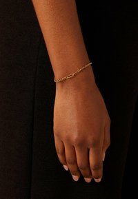 Gouden kettingarmband met vergrendelde schakels, met een delicate, gestructureerde afwerking, gedragen om een pols tegen een donkere achtergrond.