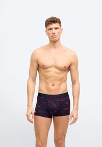 Νεαρός άνδρας όρθιος, χωρίς πουκάμισο, φορώντας μαύρα μποξεράκια με κόκκινη καρδιά και το λογότυπο Lacoste στη μέση, μπροστά σε ένα απλό φωτεινό φόντο.