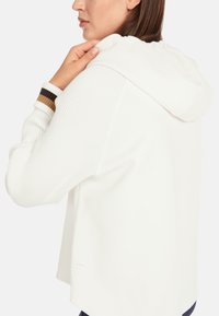 White hoodie with ribbed sleeves and a hood. Features brown and black striped accents at the cuffs. Soft texture and relaxed fit.