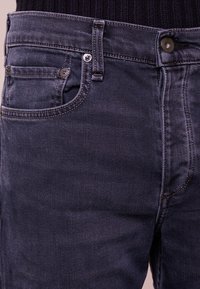 Jeans de mezclilla oscura con un corte clásico, que presentan un bolsillo delantero, costuras detalladas y botones de metal para el cierre. Textura suave.