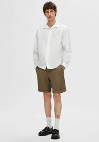 Balta marškinių su apykakle, derinama su rudo spalvos šortais. Aksesuarai apima juodus batus ir baltas kojines. Modelis stovi neutralioje aplinkoje.