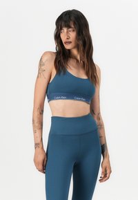 Teal sports-BH med en bred hvit logo-bånd, med en scoop-halslinje. Matchet med høyt liv teal tights, glatt tekstur.