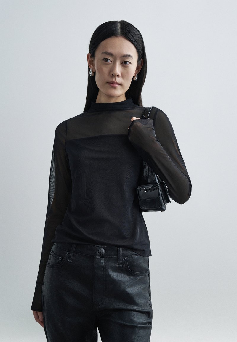 Top preto de malha de mangas compridas com gola alta, combinado com jeans pretos brilhantes. Modelo segura uma pequena bolsa preta, exibindo um design elegante.