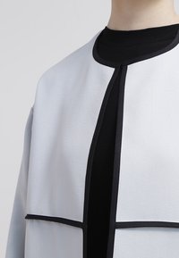 Vaaleanharmaa takki, jossa on sileä kangas, musta reunus reunassa ja jämäkkä kaula-aukko mustan paidan päällä. Ominaisuuksina minimalistinen muotoilu.