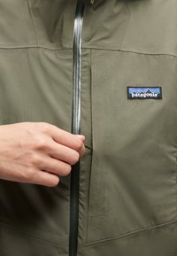 Χέρι τραβάει το φερμουάρ σε μπουφάν Patagonia ελιάς με ορατή ετικέτα λογότυπου στο αριστερό στήθος.