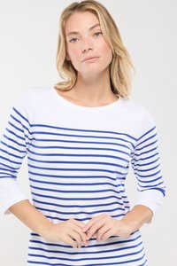 Femme blonde aux yeux bleus portant un haut à manches longues blanc avec des rayures horizontales bleues, les mains délicatement jointes devant.