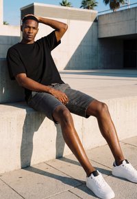 Young man in black t-shirt and gray shorts sitting on a concrete ledge outdoors, resting one hand on his head in sunlight.
