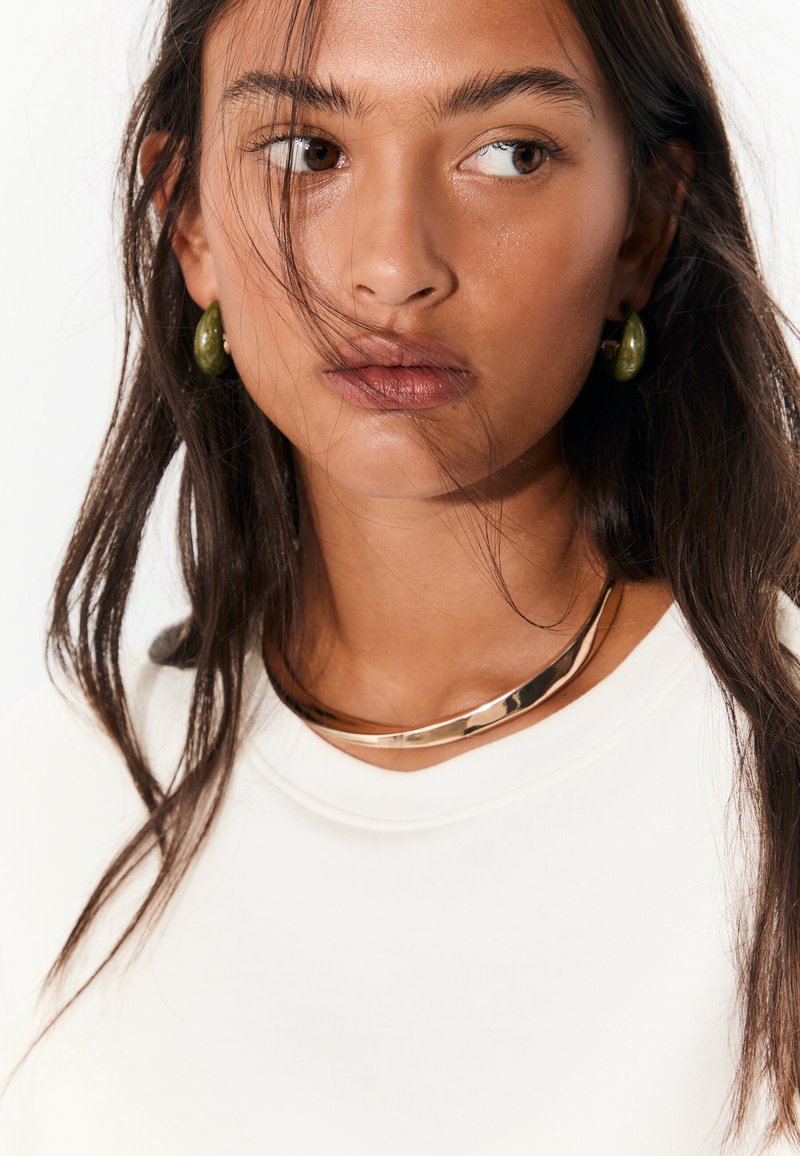 Ogrlica od metala zlatne boje, naušnice sa zelenim kamenom i kremasti top s okruglim izrezom; kosa nježno pada preko ramena.