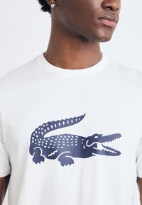 Tricou din bumbac alb, cu un grafic de crocodil de culoare albastru închis, evidențiind solzi detaliați și un design cu gura deschisă.