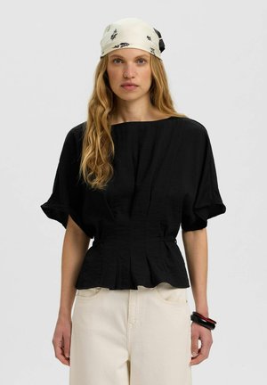 Une femme pose avec un chemisier noir à manches courtes, un pantalon crème, un foulard à motifs noué sur la tête et un bracelet foncé, sur un fond uni.
