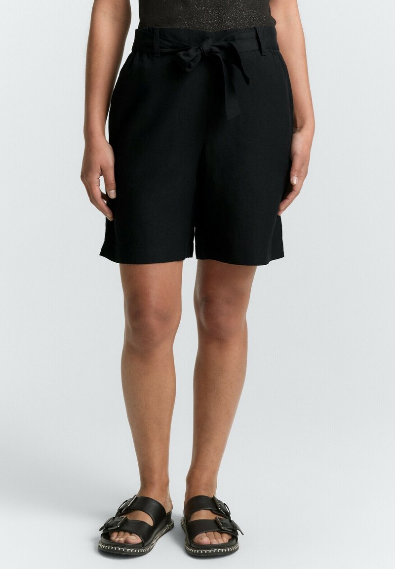 Shorts en lin noir avec une taille à nouer, présentant une coupe ample et une longueur mi-cuisse. Associés à des sandales noires à deux boucles.