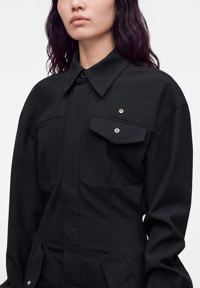 Femme portant une chemise noire boutonnée avec un col et des poches à rabat, montrée des épaules à la taille sur un fond clair uni.