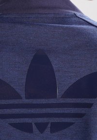 Námořnické modré polo tričko s velkým, lesklým, tmavě modrým logem ve tvaru trojlístku a třemi horizontálními pruhy na spodním okraji. Hladká textura.