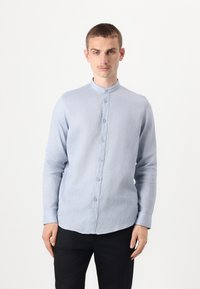 Lyseblå, langermet skjorte med mandarin krage, med knapplukking foran, laget av et teksturert stoff. Stylet med svarte bukser.