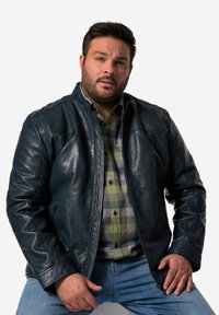 Mörkblå läderjacka med dragkedja, med stående krage och sidofickor, bärs över en grön rutig skjorta. Avslappnade denimbyxor.