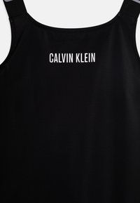 Črn top z naramnicami, ki se odlikuje po gladki teksturi in belim napisom "CALVIN KLEIN" na hrbtu, ter minimalističnem dizajnu.