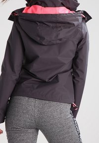 Žena v tmavě šedé bundě s růžovým lemováním a šedých texturovaných legínách s černo-bílými pruhy po stranách, otočená zády ke kameře.