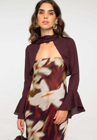 Chaqueta corta color marrón con mangas acampanadas sobre un vestido sin mangas que presenta un estampado floral difuminado en tonos blancos, marrones y rojos.
