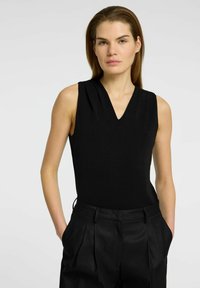 Black sleeveless top with a V-neckline, smooth texture, and pleat detailing near the shoulders; paired with high-waisted black trousers.