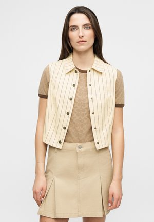 Ung kvinne iført en beige stripsvest uten ermer over en brun mønstret T-skjorte og et beige plissért skjørt, stående foran en ensfarget bakgrunn.