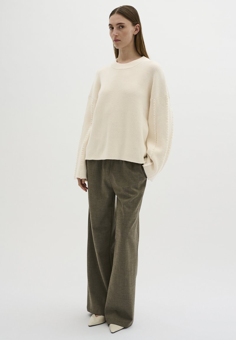 Cremefarbener, strukturierter Oversize-Pullover mit weiten Ärmeln, kombiniert mit lockeren, olivgrünen Hosen und spitzen, weißen Absatzschuhen. Neutrale Hintergrund.