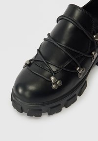 Fekete bőr cipő masszív talppal, ezüst színű fém kiegészítőkkel és vastag fekete fűzőkkel. Simább textúra, minimális varrási részletekkel.