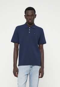 Ναυτικό μπλε πόλο μπλούζα με κλασικό γιακά, τρεις κουμπιά στο μπροστινό μέρος και μικρό λογότυπο στο στήθος; σε συνδυασμό με ανοιχτό μπλε τζιν.