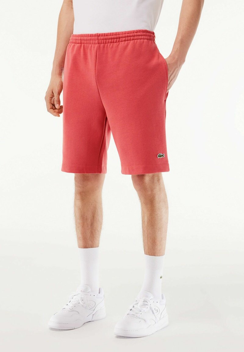 Pantalones cortos de algodón rosa para hombre con una cinturilla elástica, bolsillos laterales y un pequeño logo verde en el dobladillo. Combinados con zapatillas y calcetines blancos.