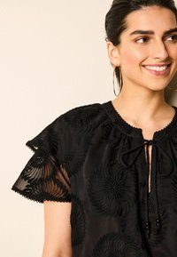 Sort broderet bluse med blomsterdesign, korte flæseærmer, en ombåndning til halsen og et let, struktureret stof.