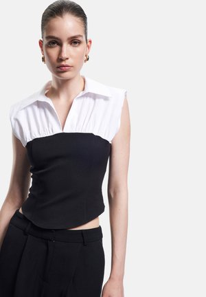 Jonge vrouw met slicked-back haar, gekleed in een mouwloze zwarte top met witte kraag en hoge zwarte broek, tegen een effen lichte achtergrond.