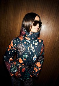 Kobieta w kolorowej kurtce z wzorami kwiatowymi i zwierzęcymi, nałożonymi dużymi, ciemnymi okularami przeciwsłonecznymi oraz w spodniach w panterkę, stojąca na tle drewnianej ściany.