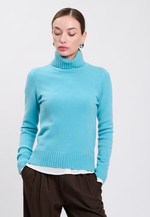 Frau trägt einen hellblauen Rollkragenpullover, darunter ein weißes Hemd, dunkelbraune Hose und silberne Creolen, die Hände in den Taschen.