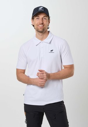 Uomo sorridente che indossa una maglietta polo bianca con un logo di alce e un berretto nero, in piedi davanti a uno sfondo chiaro e semplice.