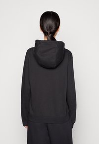 Kvinna med uppsatt hår, klädd i en svart hoodie och svarta byxor, stående mot en enkel vit bakgrund.