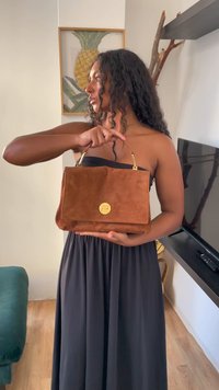 Καφέ σουέτ τσάντα χειρός με ορθογώνιο σχήμα, χρυσές λεπτομέρειες και κυκλικό λογότυπο. Η τσάντα διαθέτει δομημένο σχέδιο και ανθεκτική λαβή.