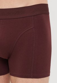 Close-up of a person wearing maroon boxer briefs with visible vertical and curved stitching and a waistband embossed with brand text.