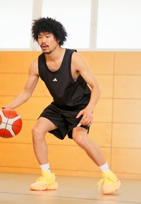 Débardeur de sport noir et shorts, chaussures de basketball jaunes vives, dribble un ballon de basketball sur un parquet intérieur avec des murs en bois.