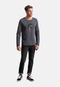 Maglietta grigia a maniche lunghe con un design grafico caratterizzato da un cerchio nero e accenti pastello, abbinata a jeans neri e sneakers nere.