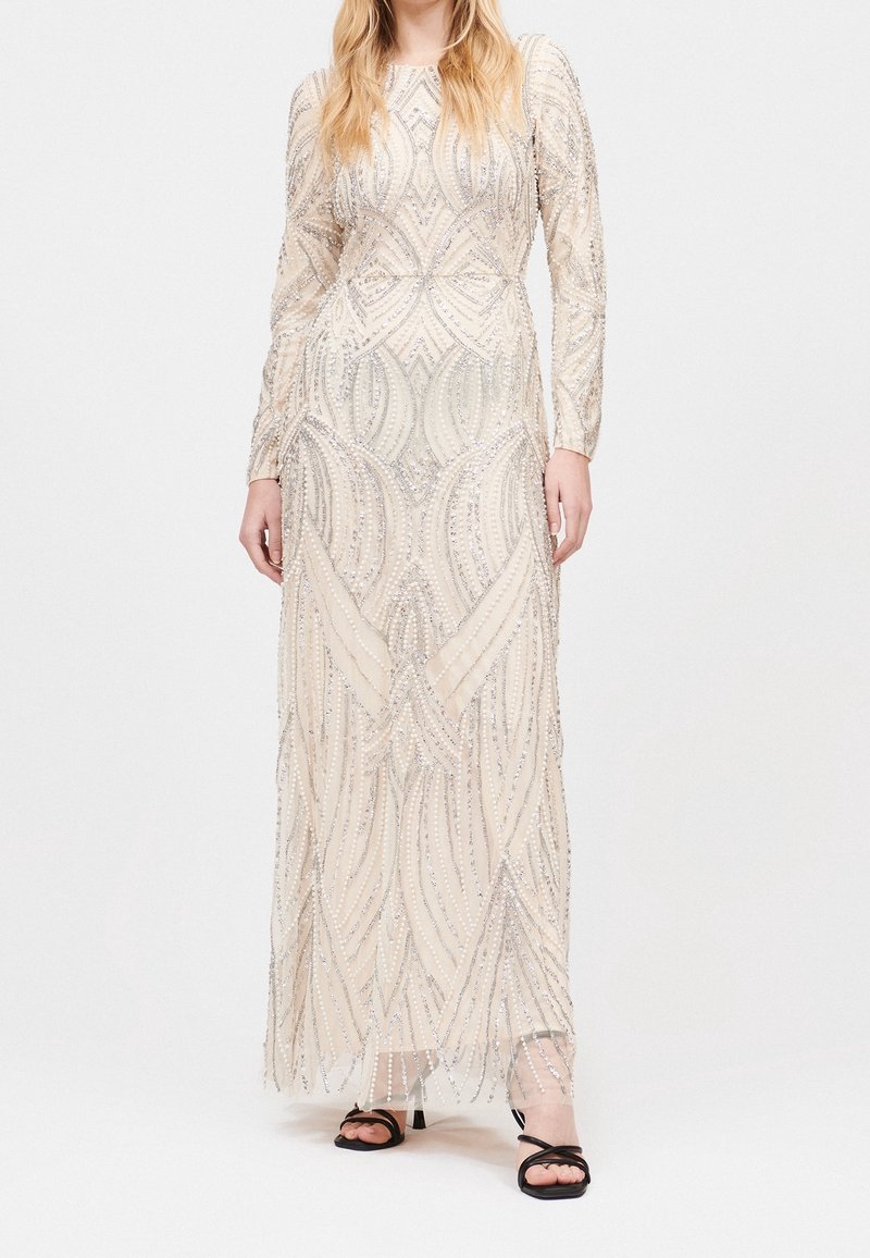 Femme portant une longue robe crème avec des motifs complexes de perles argentées et des sandales noires à talons ouverts, debout sur un fond blanc.
